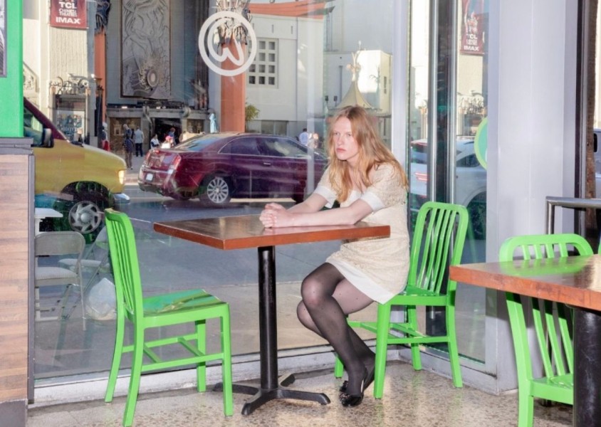 A woman with blonde hair wearing a brown dress and black tights and shoes sits at a table a table alone by a windowx